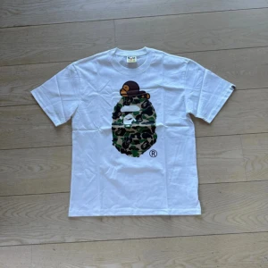 Vit t-shirt med camo-tryck från Bape - Säljer en vit t-shirt från Bape med ikoniskt camo-mönstrat ansikte och en liten apa på huvudet. Klassisk passform och rund hals. T-shirten har korta ärmar och är tillverkad i mjuk bomull. Perfekt för dig som gillar streetwear.