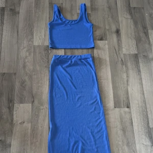 Lite mörkare blå sätt med en vacker lång kjol med slits på sidan och en fin topp - Säljer en snyggt sätt med crop top och lång kjol med ribbad struktur. Setet består av en ärmlös crop top och en lång, tight kjol med hög midja. Perfekt för dig som gillar en stilren och trendig look. Ej använd 