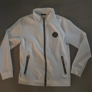 Vit kofta från Balmain med dragkedja - Säljer en snygg vit kofta från Balmain med hög krage och svart guldiga detaljer. Koftan har två fickor med dragkedja och ett Balmain-emblem på bröstet samt stor logga på ryggen. Perfekt för dig som gillar exklusiv streetwear. Nypris ligger mellan 11-12 tusen