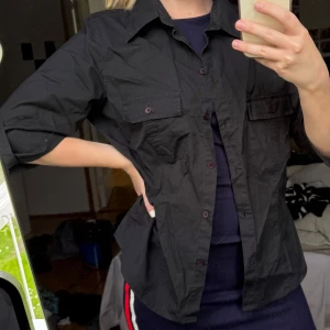 Svart skjorta med bröstfickor - Svart skjorta med klassisk krage och knäppning framtill. Två bröstfickor med lock och knappar. Skjortan har medellång ärm och är tillverkad i ett bomullsliknande material. Perfekt för en stilren och enkel look. Står storlek 44, men är mer som en S, 36