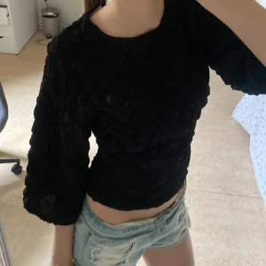 Svart blus med struktur och vida ärmar - Säljer en svart blus med coolt strukturerat tyg och trekvartsärmar. Modellen är croppad och har en lite loose passform. Perfekt för dig som gillar unika detaljer och vill sticka ut med en enkel men snygg topp.