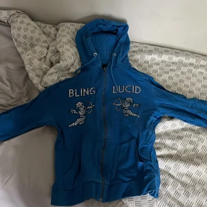 Blå hoodie med dragkedja från Bling Lucid - Säljer en blå hoodie med dragkedja och huva från Bling Lucid. Framsidan har glittrande text och ängelmotiv i strass. Perfekt för dig som vill sticka ut lite extra.