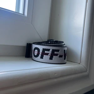 Vit och svart bälte från Off-White - Säljer ett trendigt vitt och svart bälte från Off-White med tydlig logga och svart spänne. Bältet har en industriell känsla och är perfekt för att lyfta din outfit.