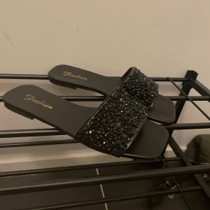 Svarta sandaler med glittriga stenar - Säljer ett par svarta sandaler med bred rem täckt av svarta glittriga stenar. Platt sula och öppen tå, perfekta för sommaren. Märkt med 'Fashion' i guld på innersulan.