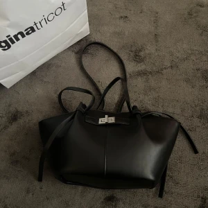 Svart handväska med silverdetalj - Snygg svart handväska från Gina Tricot med stilren silverfärgad låsdetalj framtill. Väskan har långa axelband och en mjuk, något strukturerad form. Perfekt för att bära dina viktigaste saker med stil.