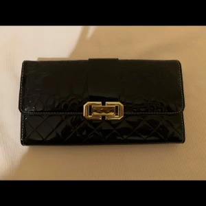 Svart plånbok med gulddetalj - Snygg svart plånbok i glansigt material med quiltad nederdel och krokopräglad ovandel. Stängs med en dekorativ guldfärgad spänne framtill. Perfekt accessoar för att hålla ordning på kort och kontanter.