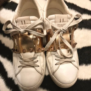 Vita Valentino Rockstud sneakers med rosé gulddetaljer - Säljer ett par vita Valentino Rockstud sneakers med rosé guldfärgade detaljer och nitar. Skorna har snörning och en klassisk rund tå. Perfekta för dig som vill ha något extra till din outfit. Fint skick. Man får med dustbag. Kan gå ner i pris via snabbaffär. Nypris ca 7500