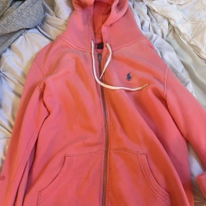 Rosa hoodie från Polo Ralph Lauren - Säljer en rosa hoodie från Polo Ralph Lauren med dragkedja och vit dragsko. Jackan har två fickor framtill och den klassiska loggan broderad på bröstet. Perfekt för en avslappnad och sportig stil.