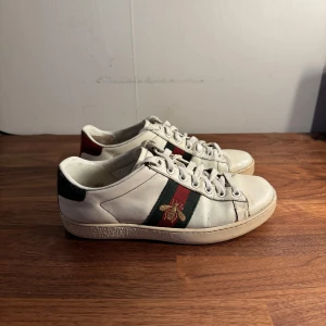 Gucci Ace sneakers - Säljer ett par vita Gucci Ace sneakers med klassiska gröna och röda ränder på sidorna samt broderad guldfärgad bi. Original pris runt 9000kr. Alla tillhör följer med och äkthetsbevis finns. Passar perfekt nu i våren och till sommaren.
