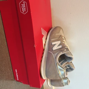Beige sneakers från New Balance - Säljer ett par snygga beige New Balance sneakers med vita detaljer och guldiga partier vid hälen. Skorna har klassisk snörning och en bekväm platt sula. Perfekta för dig som gillar stilrena och sportiga skor.