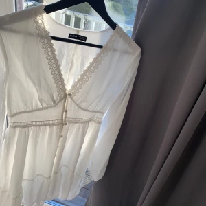 Vit långärmad klänning  - Romantisk vit klänning med långa ärmar och v-ringning, dekorerad med spets längs urringningen och knappar framtill. Lätt och luftig modell med markerad midja och volang nertill. Perfekt för vår och sommar.