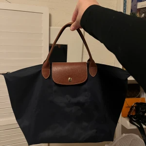 Marinblå Longchamp väska - Säljer en klassisk marinblå handväska från Longchamp med bruna skinnhandtag och lock. Väskan har silverfärgad dragkedja med Longchamp-logga och knappstängning framtill. Perfekt storlek för vardagens alla prylar. Köpt på Sellpy