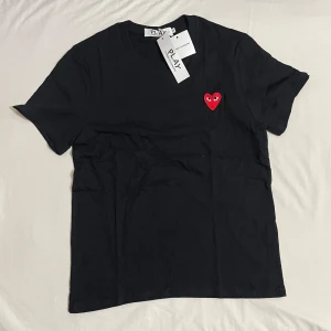 Svart t-shirt från Comme des Garçons Play - Svart t-shirt från Comme des Garçons Play med det ikoniska röda hjärtat broderat på bröstet. Klassisk passform och rund halsringning. Perfekt för en stilren och trendig look.