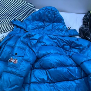 Blå pufferjacka från Moncler - Säljer en blå pufferjacka från Moncler med huva och dragkedja. Jackan har klassisk Moncler-logga på ärmen och quiltad design. Perfekt för kyliga dagar och riktigt snygg färg som sticker ut.