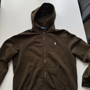 Olivgrön hoodie från Polo Ralph Lauren - Säljer en olivgrön hoodie från Polo Ralph Lauren med dragkedja och huva. Tröjan har två fickor framtill och en liten broderad logga på bröstet. Perfekt för en avslappnad stil. Färg: DARK LODEN   Nypris: 2395,00kr