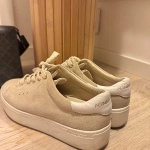 Beige sneakers från Agnes Cecilia - Säljer ett par snygga beige sneakers från Agnes Cecilia med ovandel i mocka och vit platt sula. Skorna har snörning och diskret logga på hälen och sulan. Perfekta för en stilren och avslappnad look.