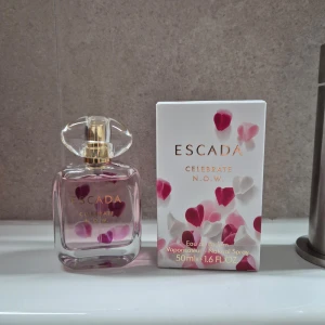 Escada Celebrate N.O.W. parfym - En söt och fräsch parfym från Escada, Celebrate N.O.W., i en genomskinlig glasflaska med guldigt lock. Flaskan har rosa detaljer och kommer med en vit kartong dekorerad med rosa och röda hjärtan. Innehåller 50 ml Eau de Parfum. Endast provsmakade.
