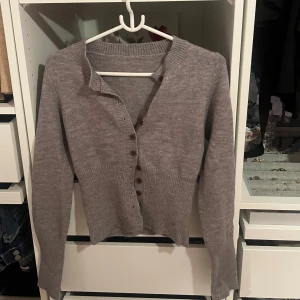 Grå stickad kofta med knappar - Säljer en grå stickad kofta med långa ärmar och knappar framtill. Koftan har ribbade muddar och en klassisk v-ringning. Passar för de som har Storlek S-M. 