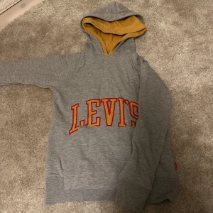 Grå hoodie från Levi's med gul huva - Säljer en grå hoodie från Levi's med stor röd och gul logga. Tröjan har en dubbel huva med gul och grå. Tröjan har långa ärmar och breda muddar vid ärmslut och nederkant. Perfekt för en avslappnad stil. 