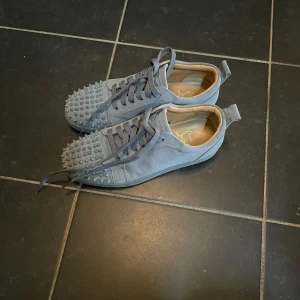 Gråa sneakers med nitar från Christian Louboutin - Säljer ett par gråa sneakers från Christian Louboutin med coola nitar framtill och klassisk röd sula. Skorna har snörning och är gjorda i mocka med rund tå. Perfekta för dig som vill sticka ut med din stil. Väldigt bra skick, storlek 43, priset kan diskuteras