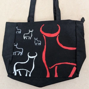 Svart tygväska med tjurmotiv - Cool svart tygväska med handtag och tryck av stiliserade tjurar i vitt och rött på framsidan. Perfekt för att bära böcker, laptop eller annat du behöver under dagen. Väskan är rymlig och har en enkel, modern design. Väskan har aldrig använts 