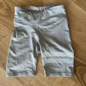 Grå/beige cykelshorts från Stronger - Grå cykelshorts från Stronger med bred resår i midjan och diskreta vita ränder med prickigt mönster på ena benet. Perfekta för träning eller en sportig look. Mjukt och stretchigt material för maximal komfort. I nyskick