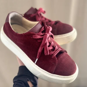 Vinröda sneakers - Säljer ett par vinröda sneakers med snörning och vit sula. Skorna har en ovandel i mocka och detaljer i skinn vid häl och plös. Perfekta för dig som vill ha en stilren och trendig look.