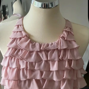 Ljusrosa volangklänning med öppen rygg - Supersöt ljusrosa klänning med flera lager volanger och öppen rygg. Klänningen har ärmlös design och halterneck, perfekt för sommarens tillfällen. Materialet känns lätt och luftigt.