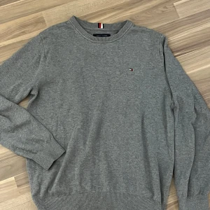 Grå sweatshirt från Tommy Hilfiger - Säljer en klassisk grå sweatshirt från Tommy Hilfiger med rund halsringning och diskret logga på bröstet. Tröjan har långa ärmar och ribbade muddar vid ärmslut och nederkant. Perfekt för en stilren och enkel look. Gjord i 100% bomull! 