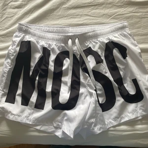 Vita badshorts med svart tryck från Moschino - Säljer ett par vita badshorts från Moschino med stora svarta bokstäver som bildar texten 'MOSCHINO' runt om. Shortsen har snörning i midjan och är tillverkade i ett lätt syntetmaterial. Perfekta för stranden eller poolen. Helt nya aldrig använda äkta moschino bad shorts