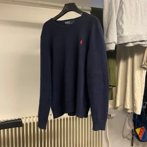 Mörkblå stickad tröja från Ralph Lauren - Mörkblå stickad tröja från Ralph Lauren med rund halsringning och diskret röd broderad logga på bröstet. Klassisk modell med ribbade muddar vid ärmslut och nederkant. Perfekt för en stilren och enkel look. Skick 8/10 nypris 2400 lite dammig nu eftersom jag ej använt den på länge.