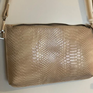 Beige axelväska i ormmönstrat skinn - Säljer en beige axelväska från Carin Wester i ormmönstrat skinn. Väskan har justerbar axelrem och stängs med dragkedja upptill. 