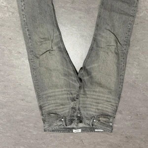Grå loose fit jeans Jack & Jones - Säljer ett par grå jeans från Jack & Jones med loose fit och knappar i gylfen. Jeansen har en klassisk femficksdesign och är i en tvättad grå nyans. Perfekta för en avslappnad stil. Jeansen har tyvärr ett veck, samt osäker på storlek (gissar på M)  därav det billiga priset!Kom gärna med bud, ifall du inte är nöjd med prissättningen💕