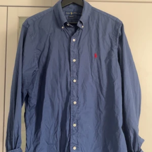 Blå skjorta från Ralph Lauren - Klassisk blå skjorta från Ralph Lauren med lång ärm och knappar framtill. Skjortan har en röd broderad logga på bröstet och button-down krage. Perfekt för en stilren och tidlös look.