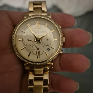 Guldig klocka från Michael Kors - Säljer en snygg guldig klocka från Michael Kors med länkarmband i metall. Klockan har romerska siffror, tre mindre urtavlor och datumvisning. Perfekt accessoar för att lyfta din stil.