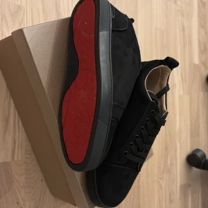 Svarta sneakers från Christian Louboutin - Säljer ett par svarta sneakers från Christian Louboutin med klassisk röd sula och snörning. Skorna har rund tå och är tillverkade i mocka med svarta detaljer. Kommer med originalkartong och dustbag. Storlek 40