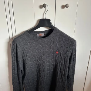 Morris tröja - Säljer en klassisk grå kabelstickad tröja från Morris i 100% merinoull. Tröjan har rund halsringning, lång ärm och en liten broderad logga i rött på bröstet. Perfekt för lager-på-lager och stilrena outfits.