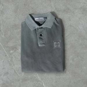 Grå pikétröja från Stone Island - Säljer en stilren grå pikétröja från Stone Island med klassisk krage och två knappar framtill. Tröjan har en diskret logotyp på bröstet och är i slim fit-modell. Perfekt för dig som gillar en clean och enkel look.