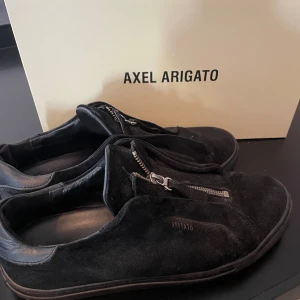 Svarta sneakers från Axel Arigato med dragkedja - Säljer ett par svarta sneakers från Axel Arigato i mocka med coola dragkedjor framtill istället för klassisk snörning. Skorna har låg profil och rund tå, perfekt för en stilren och modern look. litet hål på vänster skorn hyfsat bra skick annars pris är 500 men går att diskutera så hojta till över funderingar