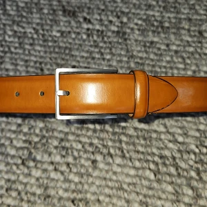 Ljusbrunt skärp från Saddler - Säljer ett ljusbrunt bälte från Saddler i äkta skinn med silverfärgat spänne. Bältet är tillverkat i Italien och har en stilren, klassisk design. Längden är 75 cm. Nypris 800kr mitt pris 249kr.  Priset är inte satt i sten. Priset är inte satt i sten