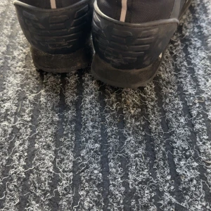 Svarta sneakers från EA7 Emporio Armani - Säljer ett par svarta sneakers från EA7 Emporio Armani med vita detaljer och logga på sidan. Skorna har snörning och är tillverkade i en kombination av syntet och textil. Perfekta för en stilren och sportig look.