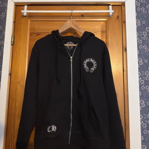 Chrome Hearts horseshoe zip hoodie - Säljer en svart hoodie från Chrome Hearts med vit logga på bröstet och stort tryck på ryggen. Ärmarna har texttryck och tröjan har dragkedja framtill samt huva med snörning. Perfekt för dig som gillar streetwear.