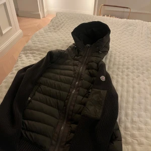 Monclear cardigan - Snygg mörk grön  pufferjacka från Moncler med stickade ärmar och huva. Jackan har dragkedja framtill, quiltad framsida och Moncler-logga på ärmen. Perfekt för kyliga dagar med även vår dagar och riktigt stilren look.