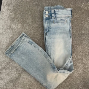 Ljusblå bootcut jeans från H&M - Säljer ett par ljusblå bootcut jeans från H&M med låg midja och justerbar midja. Jeansen har klassiska fickor fram och bak samt stängs med dragkedja och två knappar. Perfekta för en avslappnad och trendig look.
