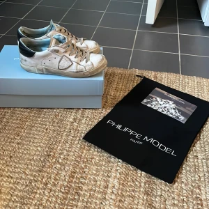 Vita sneakers från Philippe Model - Säljer ett par vita Philippe Model sneakers med grå detaljer och svart häl. Skorna har rund tå, platt sula och snörning. Klassisk design med märkets logga på sidan. Perfekta för dig som gillar stilrena och trendiga sneakers. Innersulan är lite förstörd men den går att byta. Skick 6/10. Storlek: 37 men passar 38.