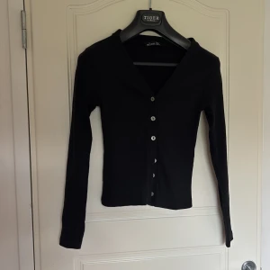 Svart ribbad kofta från Hostess - Säljer en svart ribbad kofta från Hostess i storlek S. Koftan har v-ringning, långa ärmar och knäppning framtill med silvriga knappar. Perfekt att slänga över en topp för en enkel och stilren look.