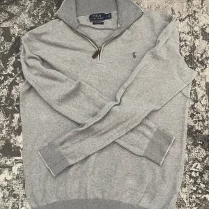 Grå halvzip tröja från Polo Ralph Lauren - Snygg grå långärmad tröja från Polo Ralph Lauren med halv dragkedja och broderad logga på bröstet. Tröjan har ribbade muddar och är perfekt för en stilren vardagslook.