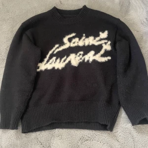 Saint Laurent stickad  - Svart stickad tröja från Saint Laurent med vit, stor logga i fuzzy stil över bröstet. Tröjan har rund halsringning och långa ärmar med ribbade muddar och nederkant. Perfekt för dig som gillar statement-plagg och exklusiva märken.