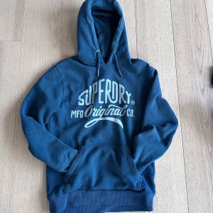 Blå hoodie från Superdry - Säljer en blå hoodie från Superdry med tryck på bröstet. Tröjan har huva med dragsko och en stor magficka. Perfekt för en avslappnad stil och passar till det mesta.