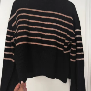 Svart stickad tröja med beige ränder - Säljer en svart stickad tröja med beige horisontella ränder. Tröjan har lång ärm och en något croppad passform. Perfekt för kyligare dagar och enkel att matcha med jeans eller kjol.